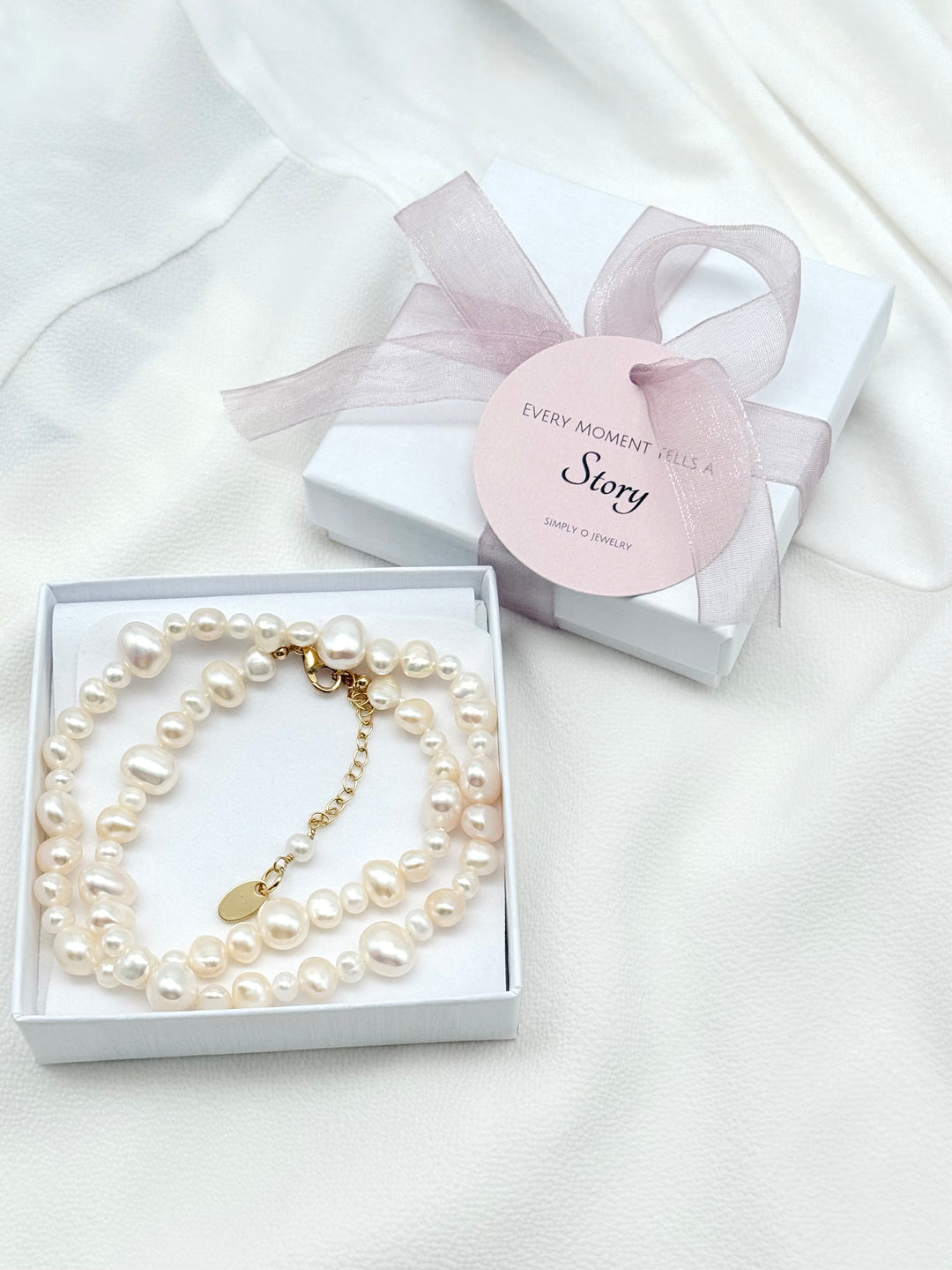 Louanne Perlenkette aus weißen Süßwasserperlen in einer Simply O Schmuckbox, daneben eine Geschenkverpackung mit roséfarbener Organzaschleife.